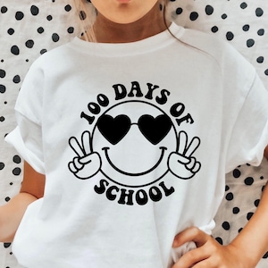 Puede incluir: Camiseta blanca con un gráfico negro de una cara sonriente con gafas de sol y signos de paz. El texto "100 Days of School" está escrito alrededor de la cara.