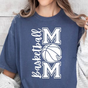 Może przedstawiać: Koszulka w kolorze granatowym z napisem "Basketball Mom" w białym kolorze. Tekst jest ułożony pionowo z grafiką koszyk&oacute;wki w środku.