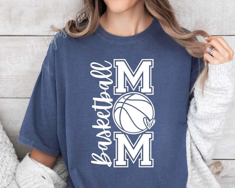 Może przedstawiać: Koszulka w kolorze granatowym z napisem "Basketball Mom" w białym kolorze. Tekst jest ułożony pionowo z grafiką koszyk&oacute;wki w środku.