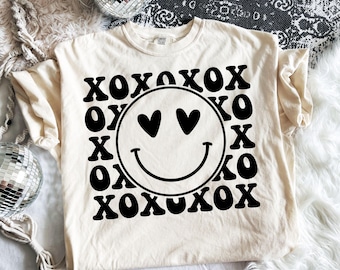 May include: A cream-colored short-sleeve t-shirt featuring a large black graphic print. The design consists of a central smiley face with heart-shaped eyes surrounded by a repeating pattern of the text XOXO in a bold, wavy black font.