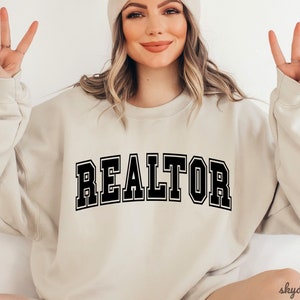 May include: A beige sweatshirt with the word "REALTOR" printed in black block letters.