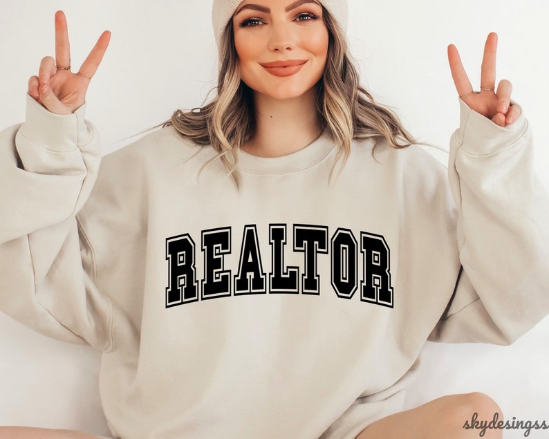 May include: A beige sweatshirt with the word "REALTOR" printed in black block letters.