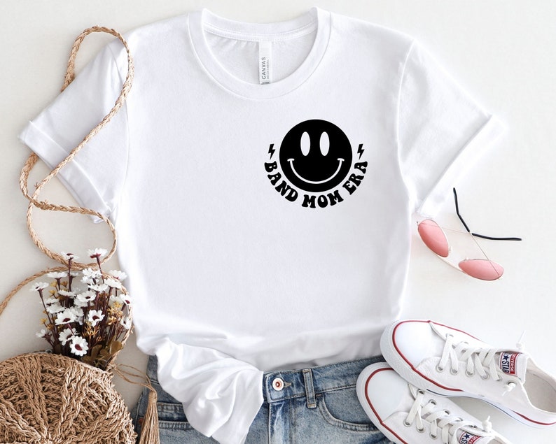 Pu&ograve; includere: Maglietta bianca con un'immagine di smiley nera e il testo "Band Mom Era" intorno.