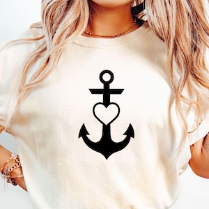 Puede incluir: Camiseta color crema con un diseño de ancla negra con un corazón en el centro. El ancla tiene una forma tradicional con una barra transversal y un anillo en la parte superior. El corazón está posicionado en el centro del ancla.