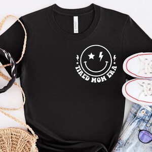 以下が含まれることがあります： 白い稲妻と「Tired Mom Era」の文字が書かれたスマイリーフェイスの白いグラフィックがプリントされた黒いTシャツ。