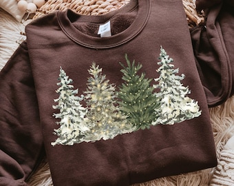 May include: A dark brown crewneck sweatshirt featuring a graphic print of four evergreen trees. The trees are depicted in shades of green and white with small yellow light accents. The sweatshirt has a ribbed collar and long sleeves.