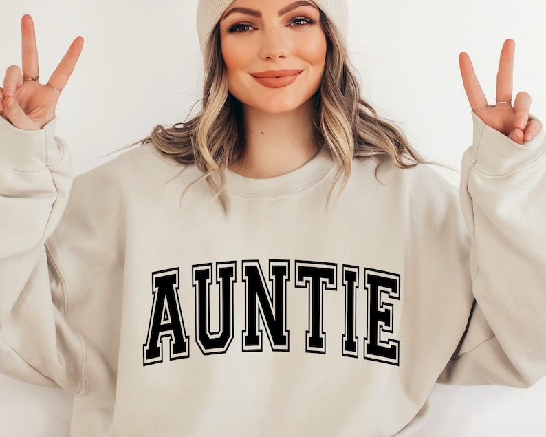 May include: A beige sweatshirt with the word "AUNTIE" printed in black block letters.