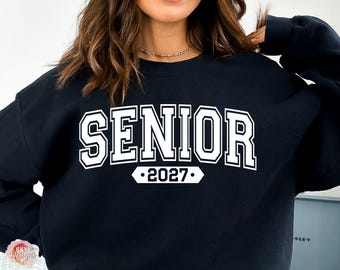 Könnte beinhalten: Ein schwarzes Sweatshirt mit Rundhalsausschnitt und dem Wort SENIOR in großen weißen Blockbuchstaben auf der Brust. Darunter befindet sich ein weißes horizontales Banner mit der Jahreszahl 2027 in schwarzer Schrift.