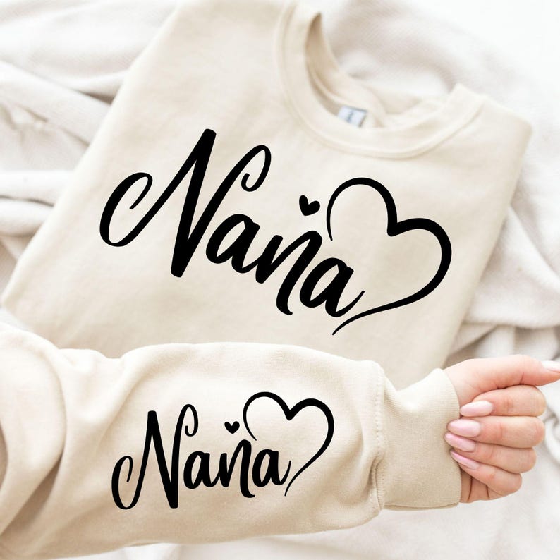Puede incluir: Una sudadera de color crema con la palabra "Naina" escrita en cursiva negra con un coraz&oacute;n al lado. La sudadera est&aacute; doblada por la mitad y la manga est&aacute; subida para mostrar el mismo dise&ntilde;o en un tama&ntilde;o m&aacute;s peque&ntilde;o.