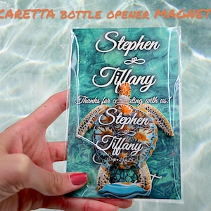 Peut inclure: Un aimant ouvre-bouteille en forme de tortue de mer avec les noms "Stephen & Tiffany" et la date "Sep. 25, 25" imprimés dessus. L'aimant est emballé dans un sac en plastique transparent sur un fond turquoise et vert.