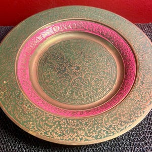 May include: Decorative round plate with an intricate floral design. The plate features a green background with gold accents and a pink inner ring. The plate's edge is also adorned with a floral pattern.