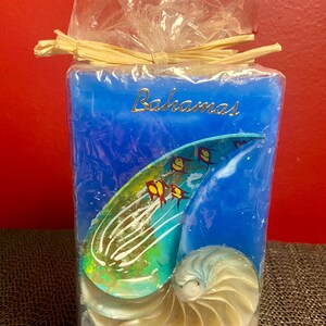 Puede incluir: Una vela azul con un diseño de concha y la palabra "Bahamas" en letras doradas. La vela presenta una concha de nautilus y peces coloridos, envuelta en celofán y atada con rafia.