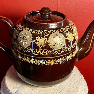 Puede incluir: Una tetera marrón con detalles decorativos dorados y blancos. La tetera tiene un cuerpo redondo y un pico curvo.