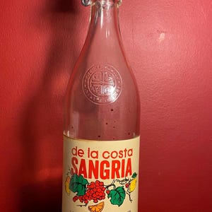 May include: A glass bottle of de la costa Sangria, a California white wine blended with natural fruit flavor. The bottle has a red and white label with a picture of fruit and the text "de la costa Sangria".