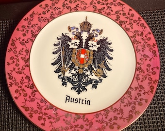 May include: A pink and gold ceramic plate with a floral design. The center of the plate features the coat of arms of Austria, with a black eagle and the text "Austria".
