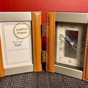 May include: A brown leather picture frame with a clock on the right side. The clock face is white with black numbers and hands. The frame is labeled "Bulova Gifts" and "Leather Frame".