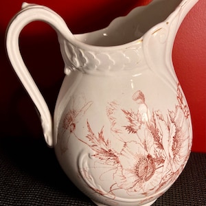 May include: White ceramic pitcher with a brown floral design. The pitcher has a wide mouth and a handle.
