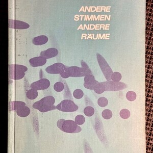 May include: A light blue book cover with the title "Andere Stimmen Andere Räume" and the author's name "Truman Capote" in white text. The cover features a design of purple abstract shapes and circles.