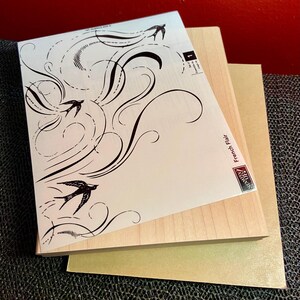 May include: A stamp set featuring a design of flying birds and swirling lines. The stamp set includes a light-colored wooden block and a paper with the words "French Flair". The image is on a dark surface.