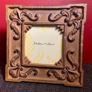 May include: Ornate brown picture frame with decorative scrollwork and a square opening. The frame has a textured finish and features the text "Milson & Louis" in the center. The background is a yellow and white checkered pattern.