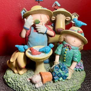 Könnte beinhalten: Eine skurrile Gartenfigur mit drei Figuren, die Strohhüte und Gartenkleidung tragen. Die Figur umfasst ein Vogelbad mit blauen und roten Vögeln, einen Hund, eine Katze und Miniatur-Vogelhäuser. Die Farbpalette umfasst Grün, Blau, Gelb und Rot.