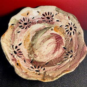 Peut inclure: Un bol en céramique avec un bord festonné et un glaçage blanc. Le bol présente un motif tourbillonnant de peinture rose, orange et verte. Le bol est décoré de motifs de fleurs découpées.