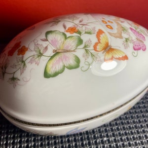 Peut inclure: Boîte à bijoux en porcelaine blanche en forme d'œuf avec une bordure dorée. Le couvercle présente un motif floral avec des papillons dans des tons de rose, orange et vert.