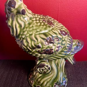 May include: A ceramic bird figurine in shades of green and brown, perched on a tree trunk base. The bird has a textured, crackled glaze finish. The figurine is a decorative item.
