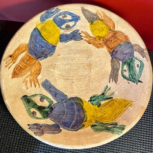 May include: A ceramic plate with a white background and three stylized frogs painted in blue, green, yellow, and brown. The frogs are facing inward towards the center of the plate.