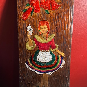 Puede incluir: Talla en relieve de madera de una mujer con un vestido rojo con un delantal blanco y una falda colorida, sosteniendo una flor. La talla está sobre un fondo marrón texturizado, con flores rojas en la parte superior.