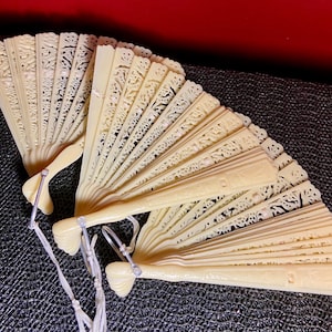 Może przedstawiać: Cztery kremowe, misternie rzeźbione wachlarze. Każdy wachlarz jest częściowo otwarty, ukazując delikatne wzory kwiatowe i geometryczne. Rączki połączone są srebrnym metalowym pierścieniem i białymi frędzlami.