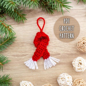 May include: A handmade red and white crochet scarf ornament with a loop for hanging. The scarf has a textured design and white fringe. The image also includes the text "PDF CROCHET PATTERN".