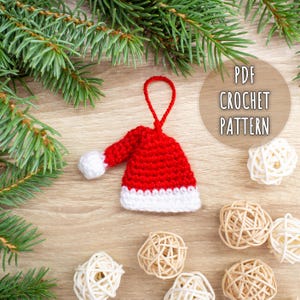 Puede incluir: Adorno de gorro de Papá Noel tejido a mano en rojo y blanco, con un lazo para colgar. El adorno se muestra sobre una superficie de madera con ramas de pino y esferas decorativas tejidas. El texto en la imagen dice "PDF CROCHET PATTERN".