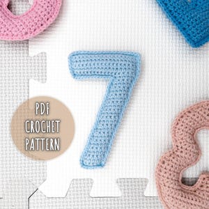 Peut inclure: Un chiffre sept crocheté bleu clair, faisant partie d'un ensemble de chiffres crochetés. L'image montre également un chiffre huit crocheté rose, un chiffre bleu crocheté et un graphique indiquant "PDF CROCHET PATTERN". Les chiffres sont sur une surface blanche.