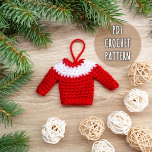 May include: A red and white crochet sweater ornament with the text "PDF CROCHET PATTERN" in a circular design. The ornament is surrounded by pine branches and decorative spheres on a wooden surface.