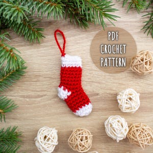 May include: A small, red and white crocheted Christmas stocking ornament with a red loop for hanging. The text "PDF CROCHET PATTERN" is in a tan circle. Decorative woven spheres and pine branches are in the background.