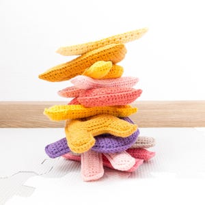 Op de afbeelding: Een stapel gehaakte banaanvormige speelgoed in verschillende tinten geel, oranje, roze en paars. Het speelgoed is gerangschikt in een piramidevorm, met de grootste banaan bovenaan en de kleinste onderaan.