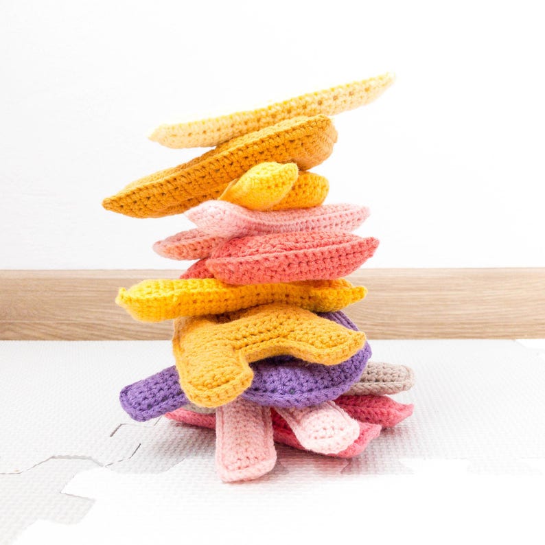 Op de afbeelding: Een stapel gehaakte banaanvormige speelgoed in verschillende tinten geel, oranje, roze en paars. Het speelgoed is gerangschikt in een piramidevorm, met de grootste banaan bovenaan en de kleinste onderaan.