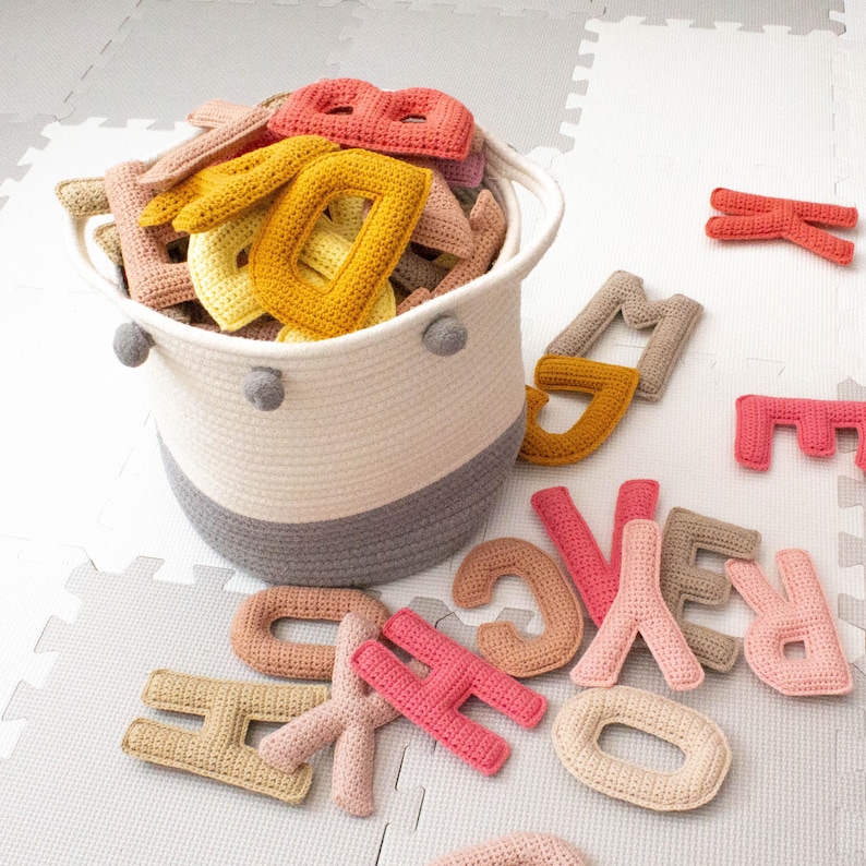 Op de afbeelding: Een geweven mand gevuld met gehaakte letters in verschillende kleuren, waaronder geel, oranje, roze en bruin. De letters zijn verspreid rond de mand op een witte en grijze betegelde vloer.
