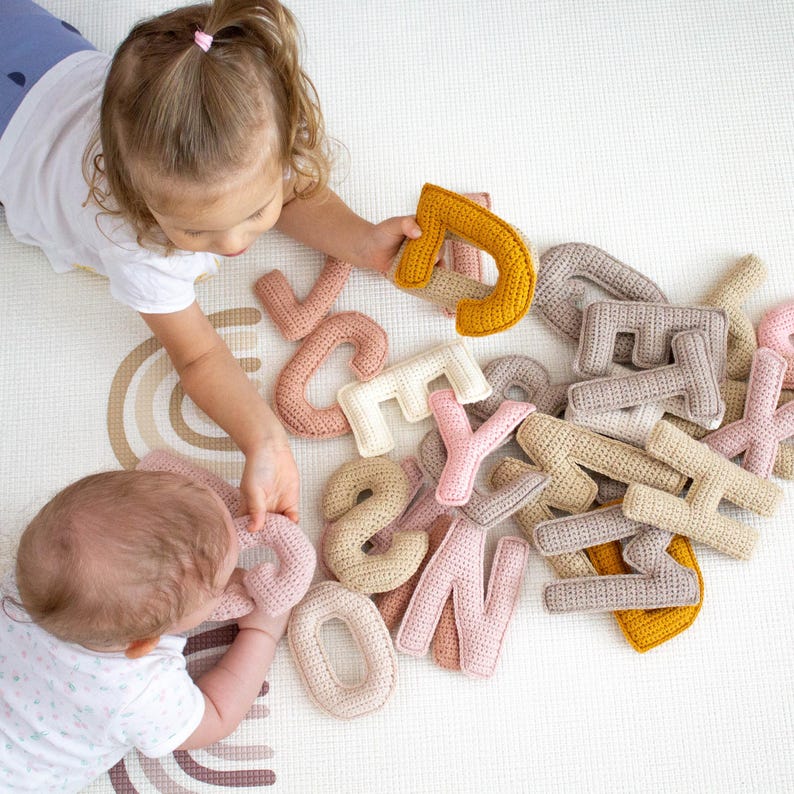 Op de afbeelding: Twee kinderen spelen met een set gehaakte alfabetletters in verschillende kleuren, waaronder beige, roze en geel. De letters zijn zacht en knuffelig, perfect voor kleine handjes om te verkennen.
