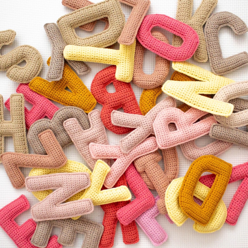 Op de afbeelding: Een verzameling gehaakte letters in verschillende kleuren, waaronder roze, geel, oranje, bruin en wit. De letters zijn in een willekeurig patroon gerangschikt, waardoor een kleurrijk en speels display ontstaat.
