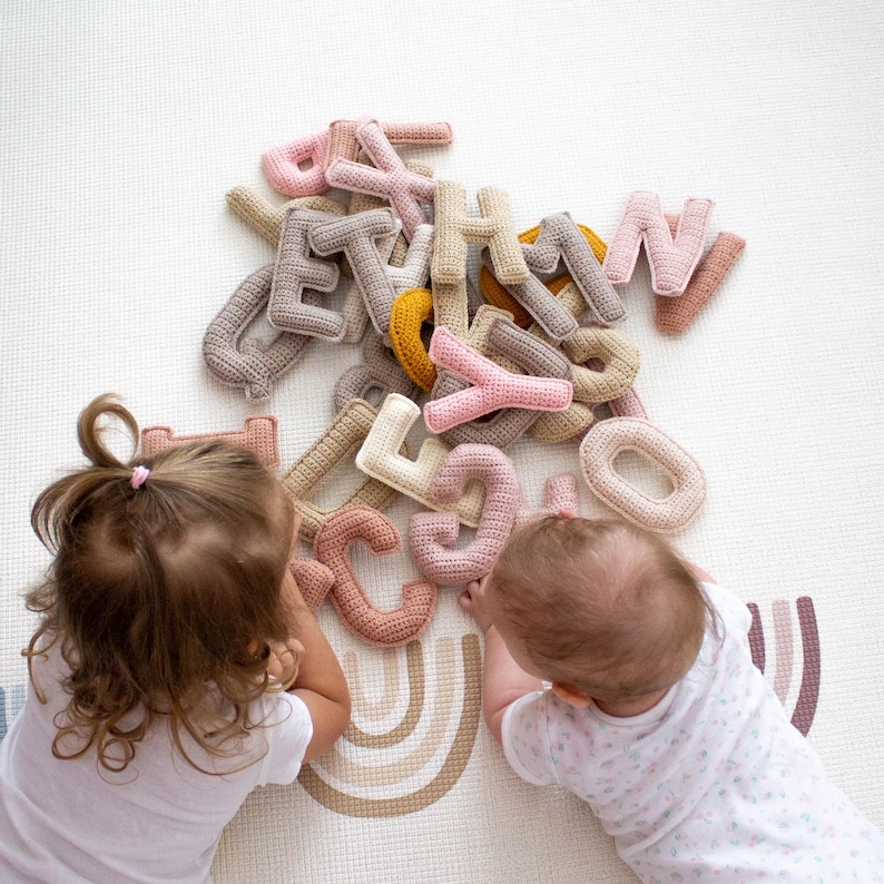 Op de afbeelding: Twee baby's spelen met een stapel gehaakte alfabetletters in verschillende kleuren, waaronder roze, beige en geel. De letters liggen verspreid op een wit en beige kleed met een regenboogpatroon.