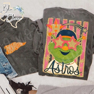 Puede incluir: Dos camisetas de manga larga de color gris oscuro. Una presenta la palabra "Houston" en naranja. La otra muestra un personaje de dibujos animados verde con gafas de sol en forma de estrella, con la palabra "Astros" debajo. Las camisetas están sobre una superficie blanca.