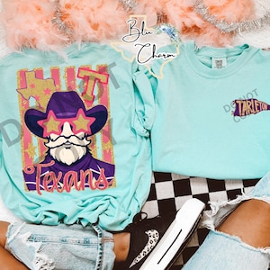 May include: A light turquoise t-shirt featuring a graphic of a cowboy with a beard and star-shaped glasses, the word Texans, and a Texas state outline. The front chest area displays a small purple pennant with the text Tarleton printed on it.
