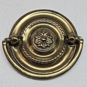 Puede incluir: Un tirador de cajón de latón con un diseño floral central. El tirador tiene forma circular con una textura de cuerda alrededor del centro y un asa a cada lado. El herraje es probablemente para muebles.