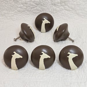May include: Six brown cabinet knobs featuring a cream-coloured giraffe design. The knobs are round with a curved profile and a metal screw for fitting. The giraffe design is a simple, stylized silhouette.