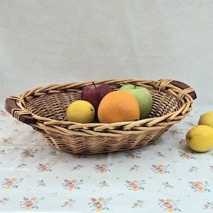 May include: Oval woven wicker basket with wooden handles, filled with fresh fruit. The basket contains a red apple, a green apple, an orange, and a lemon. Additional lemons are scattered nearby. The basket sits on a floral patterned tablecloth.