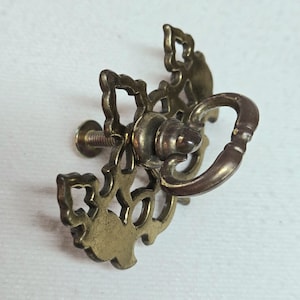 May include: Ornate brass drawer pull with a decorative openwork design and a curved handle. The hardware features a screw for easy installation and adds a vintage touch to furniture. Ideal for cabinets and drawers.