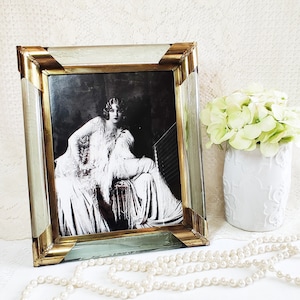 May include: A framed black and white photograph of a woman in a vintage style dress. The frame is gold and light green. A white vase with green flowers and a pearl necklace are also in the image.