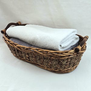 May include: A woven wicker basket with dark wooden handles, filled with folded white and gray towels. The basket is oval-shaped and sits on a white surface, suggesting a bathroom or spa setting.